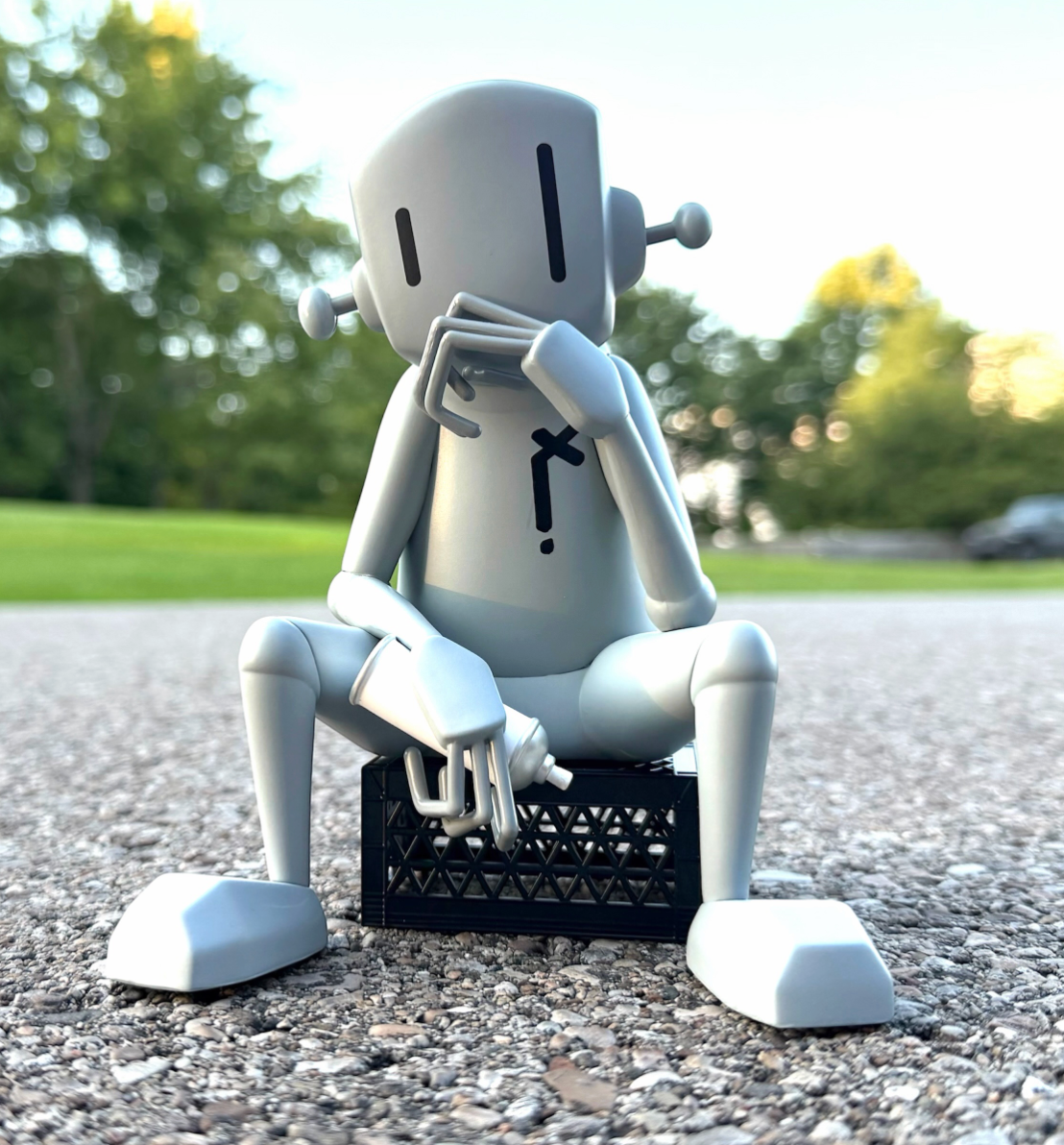 A light gray robot figurine sitting on a black crate outdoors on a gravel path with blurred green trees in the background.